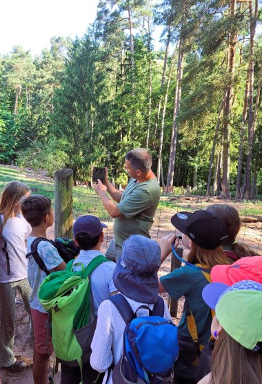 Die Freie Bauernhof-Waldschule Südpfalz in Völkersweiler hat bei einer Mitmachaktion mit einem Informationsschild über Wildschweine einen Rundgang mit Parkranger Daniel Kraus durch den Wild- und Wanderpark bei Silz gewonnen. Foto: WWP Kinder schauen zu Daniel Kraus, der die Form eines Wildschwein-Fußabdrucks hochhält.