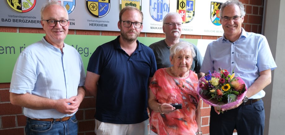 Fünf Personen stehen vor einer Wappenwand, die Frau vorne hält eine Ehrennadel.