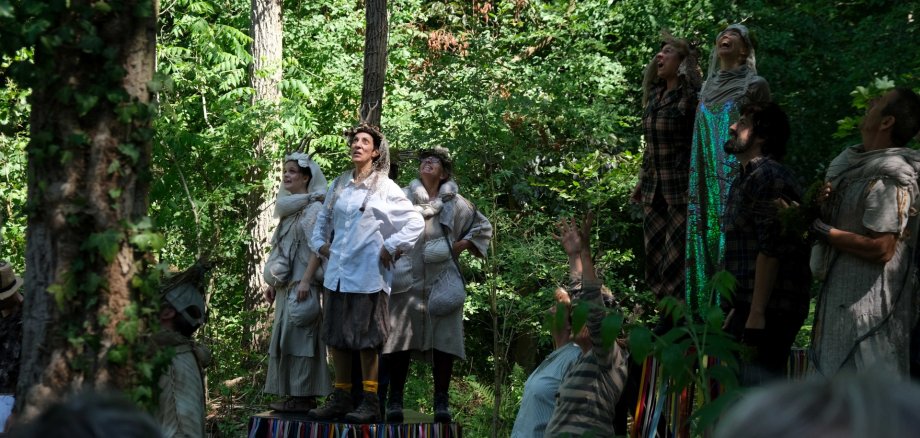 Szene aus „Das Grüne Herz“. Foto: KV SÜW Mehrere Darstellerinnen und Darsteller in fantasievollen Kostümen stehen während einer Freiluftaufführung in einem Wald. Einige Personen blicken nach oben, andere heben die Arme oder gestikulieren. Die Szene spielt zwischen hohen Bäumen und dichtem Grün, während im Vordergrund Zuschauerinnen und Zuschauer zu erkennen sind.