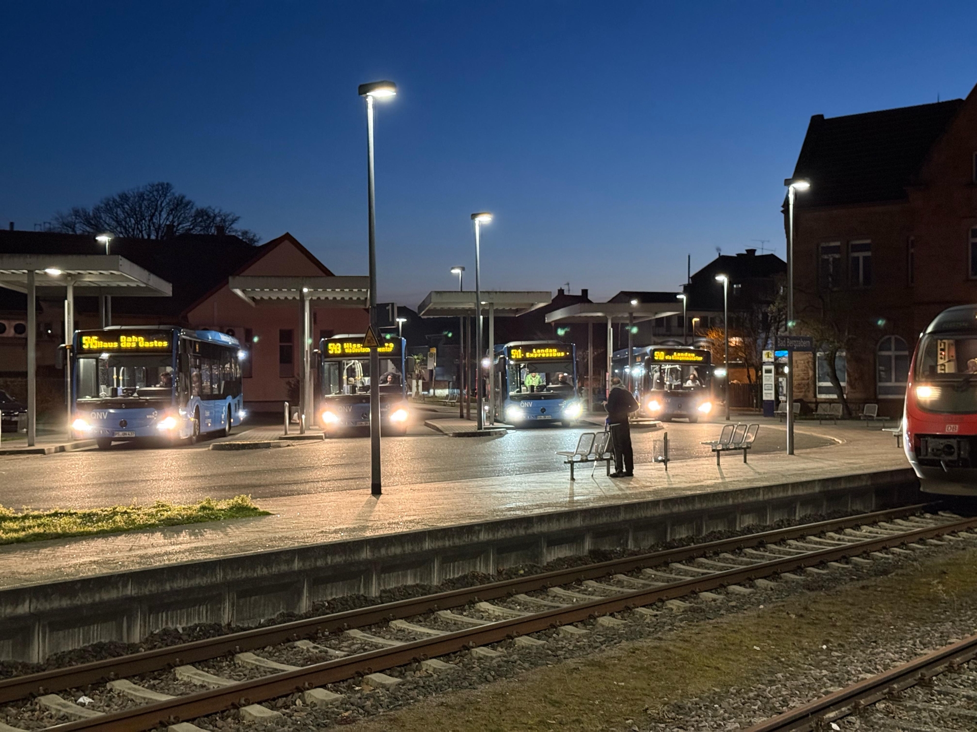 Mehrere Linienbusse stehen in den Abendstunden an den Bussteigen eines Busbahnhofs neben einem Bahnhofsgelände. Im Vordergrund verlaufen Bahngleise, rechts ist ein Teil eines Regionalzugs zu sehen. Die Busse mit beleuchteten Zielanzeigen warten unter modernen Überdachungen, während einzelne Personen auf dem Bahnsteig stehen. Straßenlaternen erhellen die Szene vor dem dunkelblauen Himmel.