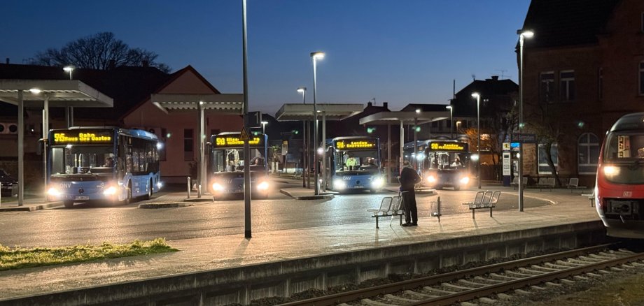 Mehrere Linienbusse stehen in den Abendstunden an den Bussteigen eines Busbahnhofs neben einem Bahnhofsgelände. Im Vordergrund verlaufen Bahngleise, rechts ist ein Teil eines Regionalzugs zu sehen. Die Busse mit beleuchteten Zielanzeigen warten unter modernen Überdachungen, während einzelne Personen auf dem Bahnsteig stehen. Straßenlaternen erhellen die Szene vor dem dunkelblauen Himmel.