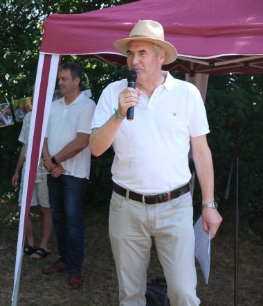 Landrat Dietmar Seefeldt spricht bei einer Freiluftveranstaltung zu den Gästen. Er trägt einen hellen Sonnenhut und hält ein Mikrofon sowie Unterlagen in der Hand.