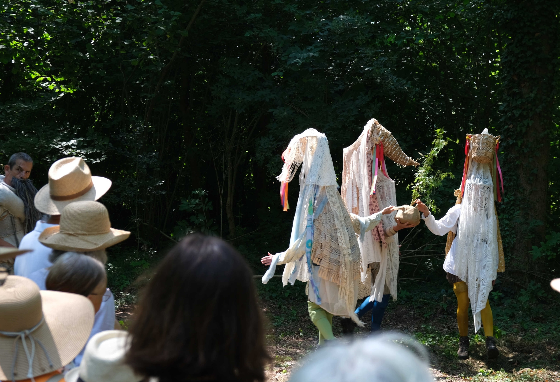 Schauspielerinnen und Schauspieler in fantasievollen Kostümen treten während des Theaterspaziergangs im Wald auf. Im Vordergrund verfolgen Besucherinnen und Besucher die Aufführung, im Hintergrund ist dichter Baumbestand zu sehen.
