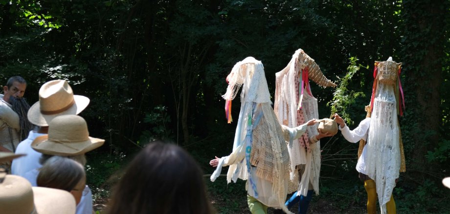 Schauspielerinnen und Schauspieler in fantasievollen Kostümen treten während des Theaterspaziergangs im Wald auf. Im Vordergrund verfolgen Besucherinnen und Besucher die Aufführung, im Hintergrund ist dichter Baumbestand zu sehen.