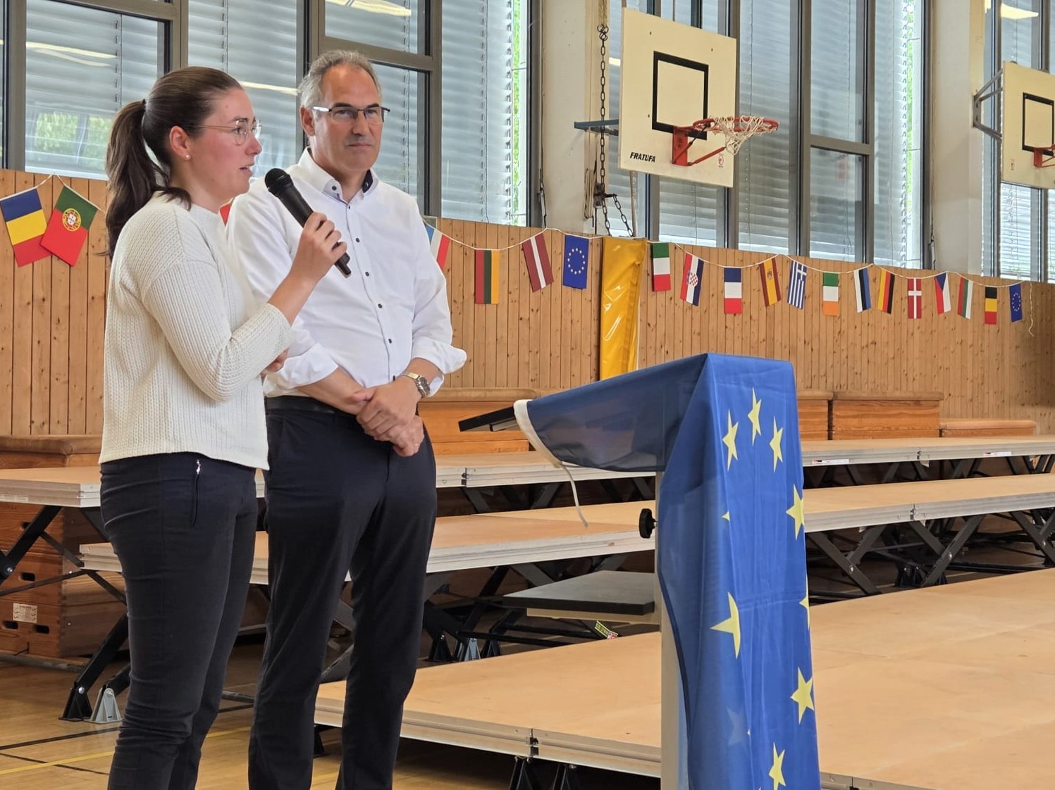 Eine Frau spricht mit einem Mikrofon in einer Sporthalle, während ein Mann neben ihr steht. Vor ihnen befindet sich ein Rednerpult, das mit einer Europa-Flagge bedeckt ist.