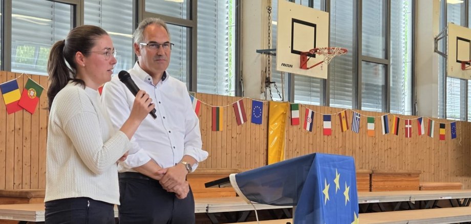 Eine Frau spricht mit einem Mikrofon in einer Sporthalle, während ein Mann neben ihr steht. Vor ihnen befindet sich ein Rednerpult, das mit einer Europa-Flagge bedeckt ist.