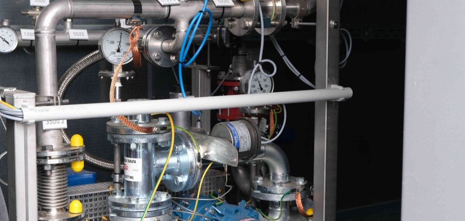 Eine Schwachgasanlage wird auf der ehemaligen Hausmülldeponie Edesheim errichtet werden, hier ein Foto von einer Komponente der bereits bestehenden Anlage auf der früheren Deponie in Heuchelheim. Foto: KV SÜW Blick in eine technische Anlage mit Rohrleitungen, Ventilen, Messgeräten und einer blauen Pumpe. Mehrere Druckanzeigen und gekennzeichnete Armaturen sind an den metallischen Leitungen angebracht. Die Installation befindet sich in einem geschlossenen Technikraum und dient der Erfassung und Steuerung von Gasströmen.