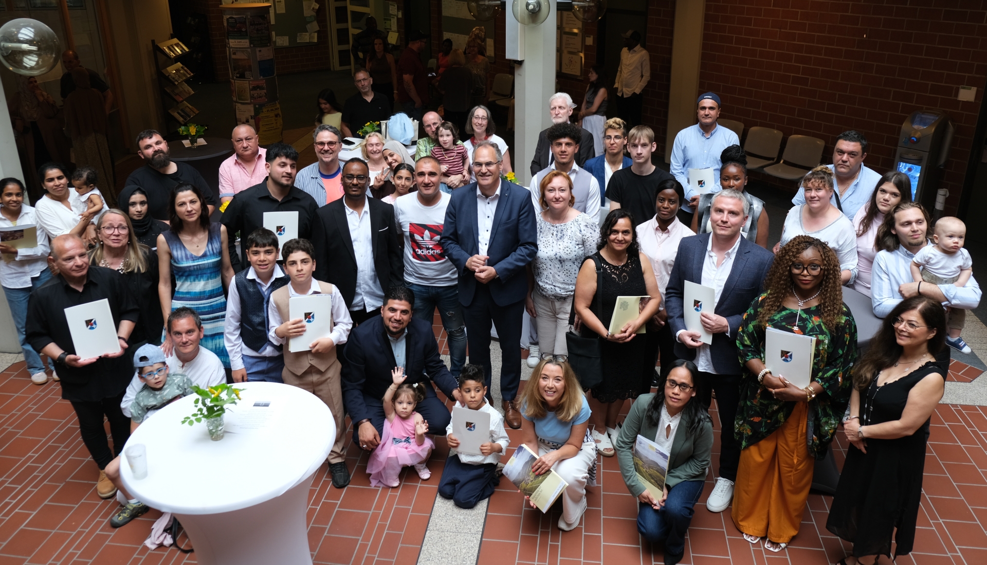 Gruppenfoto einer Einbürgerungsfeier im Foyer des Kreishauses Südliche Weinstraße. Rund 50 Erwachsene und Kinder unterschiedlicher Herkunft stehen und sitzen gemeinsam für das Foto, viele halten Einbürgerungsurkunden oder Mappen in den Händen. Im Mittelpunkt der Gruppe steht Landrat Dietmar Seefeldt. Die Aufnahme vermittelt die feierliche Atmosphäre der Veranstaltung.