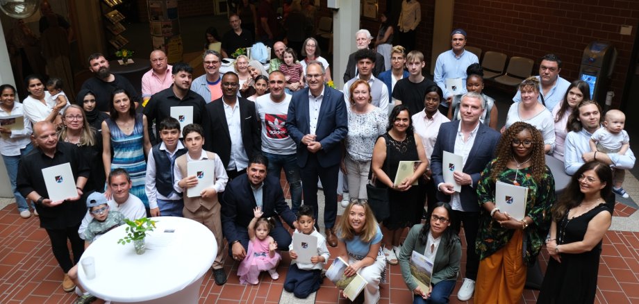 Nach der feierlichen Einbürgerungsfeier im Kreishaus SÜW. Foto: KV SÜW Gruppenfoto einer Einbürgerungsfeier im Foyer des Kreishauses Südliche Weinstraße. Rund 50 Erwachsene und Kinder unterschiedlicher Herkunft stehen und sitzen gemeinsam für das Foto, viele halten Einbürgerungsurkunden oder Mappen in den Händen. Im Mittelpunkt der Gruppe steht Landrat Dietmar Seefeldt. Die Aufnahme vermittelt die feierliche Atmosphäre der Veranstaltung.
