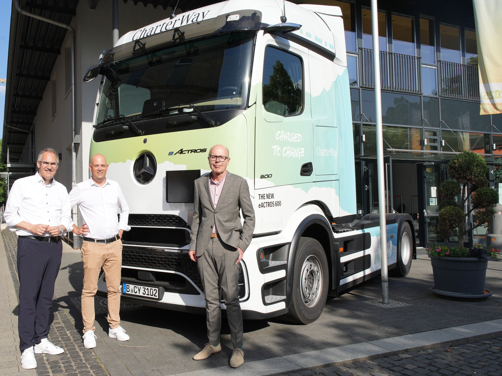 Drei Männer stehen vor einer modernen Glasfassade neben einer weißen elektrischen Sattelzugmaschine vom Typ Mercedes-Benz eActros 600. Das Fahrzeug trägt die Aufschrift „CharterWay“ und ist vor dem Eingangsbereich eines Gebäudes geparkt.