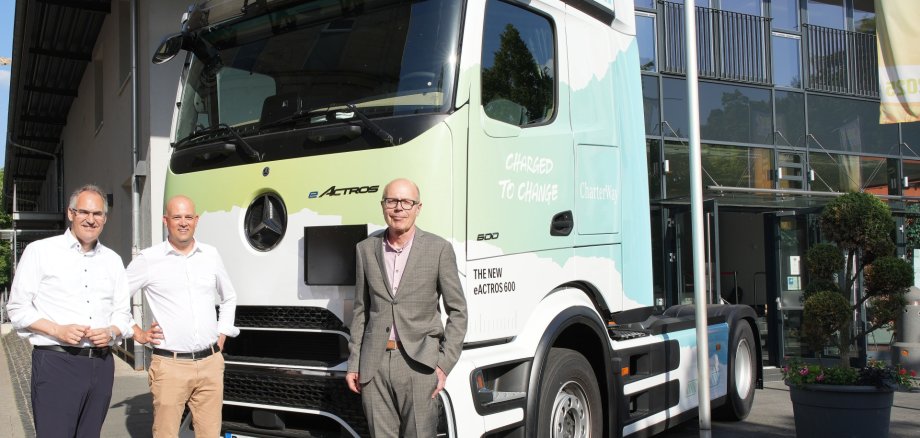 Forum Ladeinfrastruktur für die Logistik:
Südpfalz setzt Impulse für die Zukunft des elektrischen Güterverkehrs. Foto: KV GER Drei Männer stehen vor einer modernen Glasfassade neben einer weißen elektrischen Sattelzugmaschine vom Typ Mercedes-Benz eActros 600. Das Fahrzeug trägt die Aufschrift „CharterWay“ und ist vor dem Eingangsbereich eines Gebäudes geparkt.