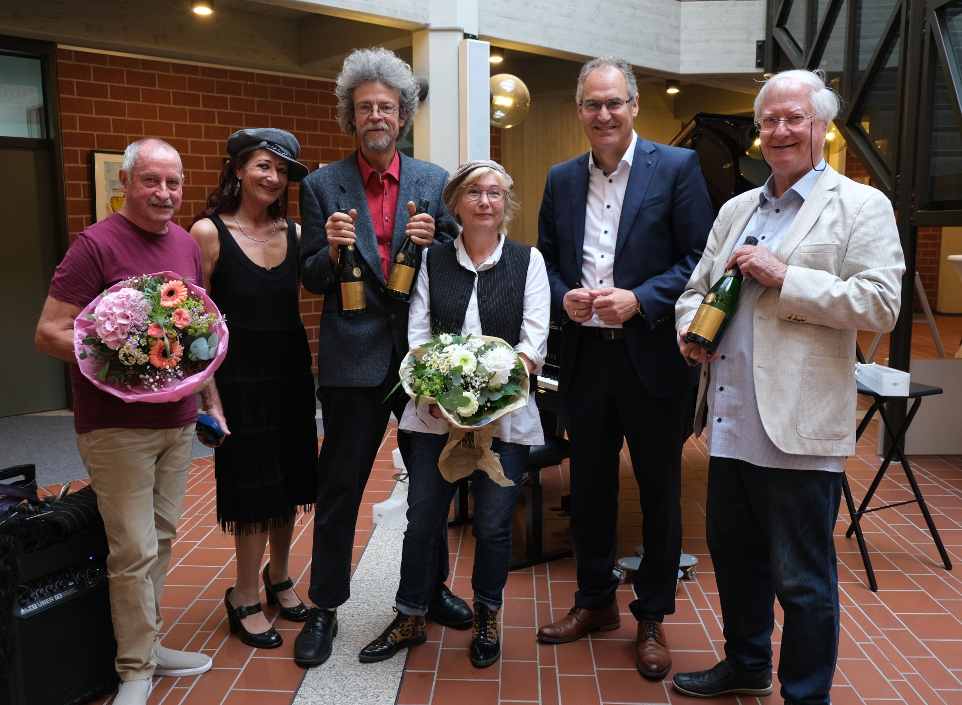 Sechs Personen stehen gemeinsam für ein Foto im Foyer eines Gebäudes. Zwei Personen halten Blumensträ0ße in den Händen, drei weitere präsentieren Sektflaschen als Geschenk.