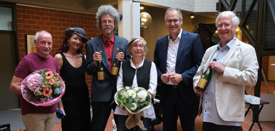 Sechs Personen stehen gemeinsam für ein Foto im Foyer eines Gebäudes. Zwei Personen halten Blumensträ0ße in den Händen, drei weitere präsentieren Sektflaschen als Geschenk.