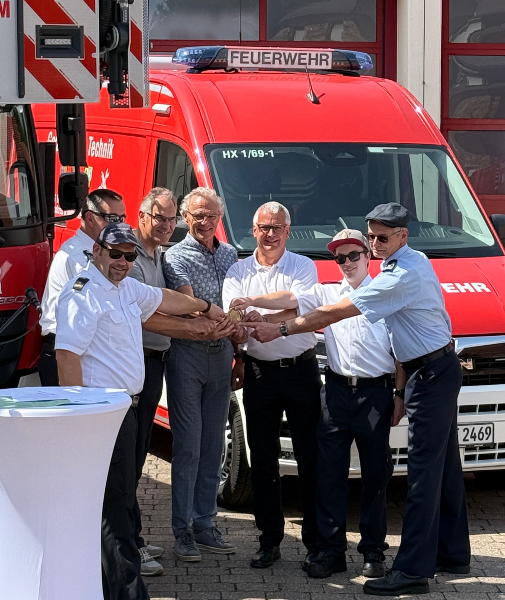 Mehrere Personen stehen vor einem roten Feuerwehrfahrzeug und legen symbolisch ihre Hände übereinander. Unter ihnen befinden sich Feuerwehrangehörige in Uniform sowie weitere Beteiligte in Alltagskleidung.