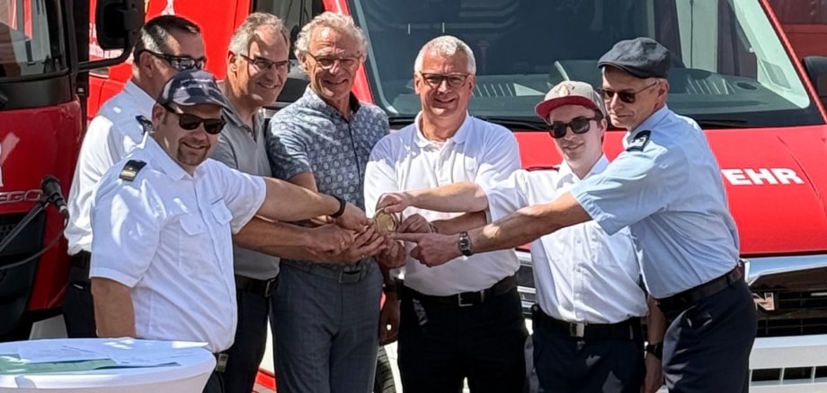 Schlüsselübergabe für den Gerätewagen „Technik“ (von links): Jens Thiele, Brand- und Katastrophenschutzinspekteur der Südlichen Weinstraße, Wehrleiter der VG Herxheim Jürgen Fink, Landrat Dietmar Seefeldt, die Bürgermeister Axel Wassyl (VG Offenbach) und Christian Sommer (VG Herxheim), Gerätewart Ari Gadinger und der stellvertretende Wehrleiter der VG Offenbach Joachim Wagner freuen sich über die gelungene Kooperation. Foto: Feuerwehr Herxheim Mehrere Personen stehen vor einem roten Feuerwehrfahrzeug und legen symbolisch ihre Hände übereinander. Unter ihnen befinden sich Feuerwehrangehörige in Uniform sowie weitere Beteiligte in Alltagskleidung.