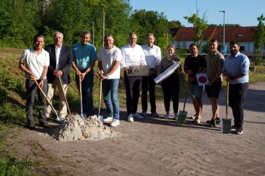 Zehn Personen stehen bei einem symbolischen Spatenstich auf einer freien Fläche neben jungen Bäumen und halten Spaten in den Händen. Im Hintergrund sind Wohnhäuser und ein Waldrand zu sehen. Vor der Gruppe liegt ein kleiner Erdhaufen, der den Beginn eines Bauprojekts markiert.