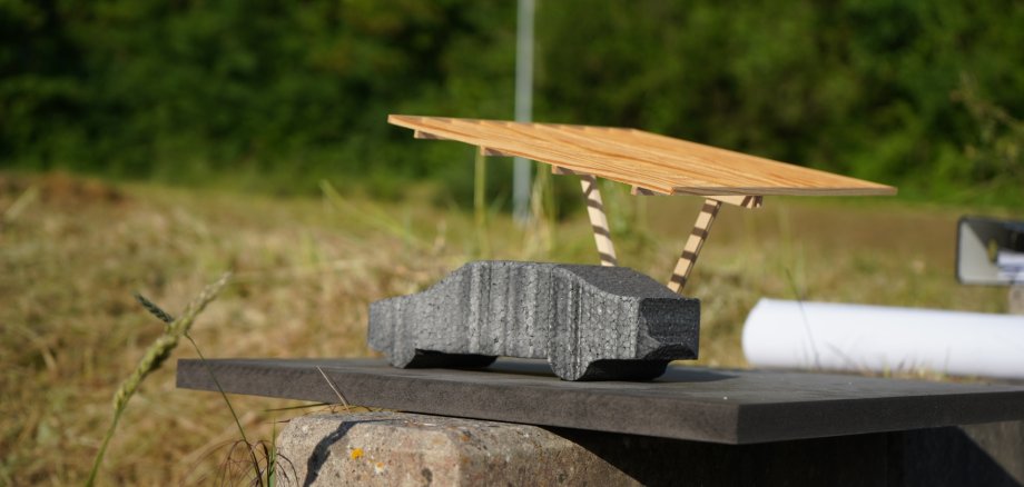 Ein kleines Architekturmodell einer Parkplatzüberdachung mit Solarmodulen steht auf einem Steinsockel. Das Modell zeigt eine dunkle Stellfläche mit einer filigranen Holzkonstruktion und einem geneigten Dach. Im Hintergrund sind Wiese und Bäume unscharf zu erkennen.
