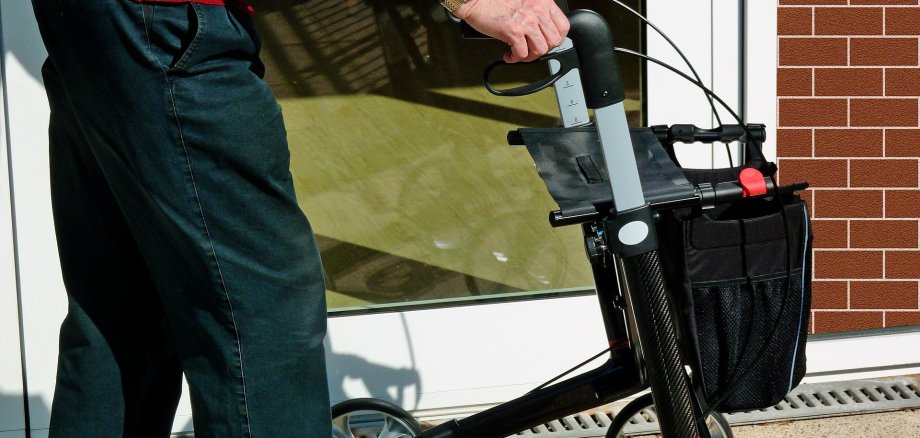 Beine und ein Rollator vor einer Fensterfront.