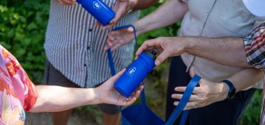 Mehrere Personen reichen sich blaue Trinkflaschen und Stofftaschen mit gleichem Logo. Zu sehen sind nur die Hände und Oberkörper der Beteiligten. Die Szene spielt im Freien vor grünem Hintergrund und vermittelt den Eindruck einer gemeinsamen Aktion zur Ausgabe von wiederverwendbaren Trinkflaschen.