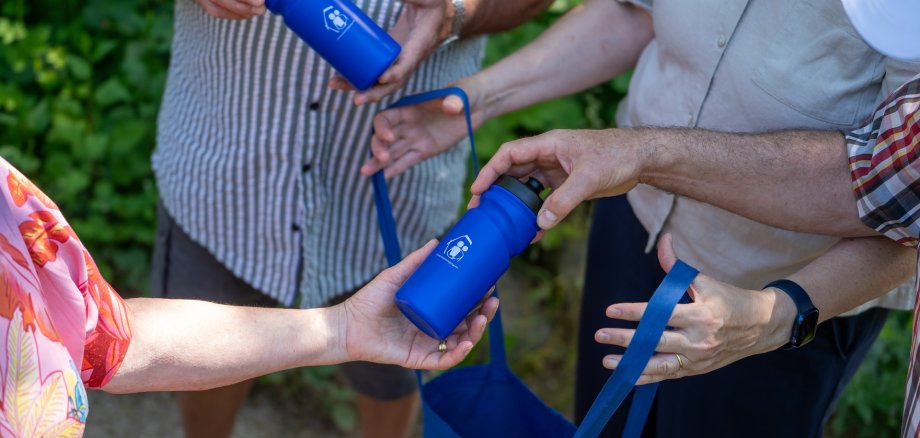 Die Fachkräfte Gemeindeschwester plus im Kreis SÜW raten Seniorinnen und Senioren dazu, während der heißen Tage viel zu trinken. Passend dazu verteilen sie bei ihren Hausbesuchen und den Plauderspaziergängen Trinkflaschen. Foto: KV SÜW Mehrere Personen reichen sich blaue Trinkflaschen und Stofftaschen mit gleichem Logo. Zu sehen sind nur die Hände und Oberkörper der Beteiligten. Die Szene spielt im Freien vor grünem Hintergrund und vermittelt den Eindruck einer gemeinsamen Aktion zur Ausgabe von wiederverwendbaren Trinkflaschen.