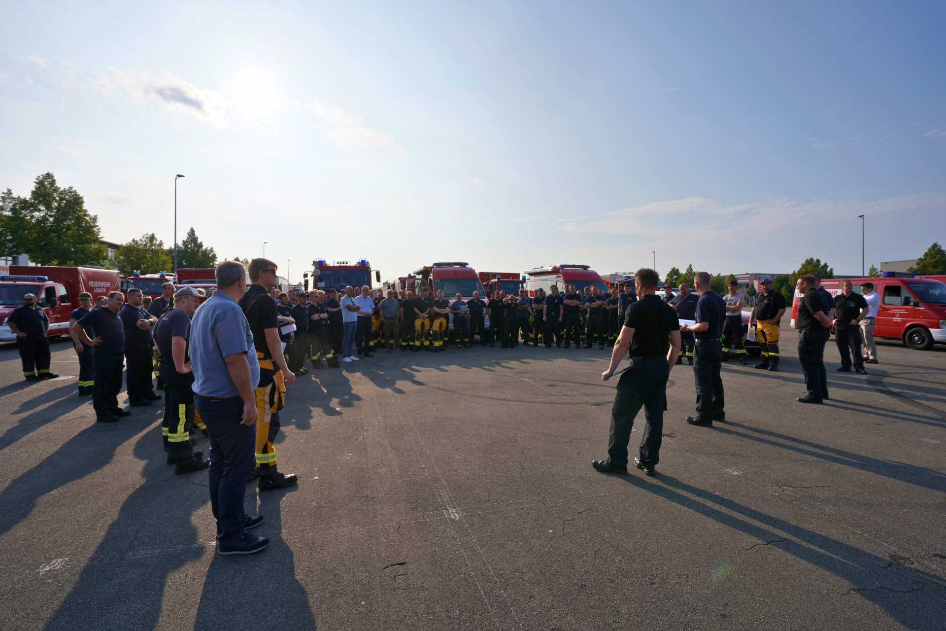 Einsatzkräfte des Brand- und Katastrophenschutzes aus dem Bereich der integrierten Leitstelle Landau versammeln sich mit ihren Fahrzeugen auf dem Neuen Messplatz in Landau vor dem gemeinsamen Abmarsch zu einem Waldbrandeinsatz. Eine Person spricht zur Gruppe.
