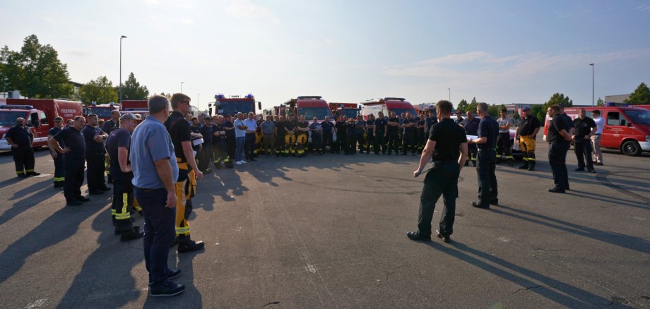 Einsatzkräfte des Brand- und Katastrophenschutzes aus dem Bereich der integrierten Leitstelle Landau versammeln sich mit ihren Fahrzeugen auf dem Neuen Messplatz in Landau vor dem gemeinsamen Abmarsch zu einem Waldbrandeinsatz. Eine Person spricht zur Gruppe.