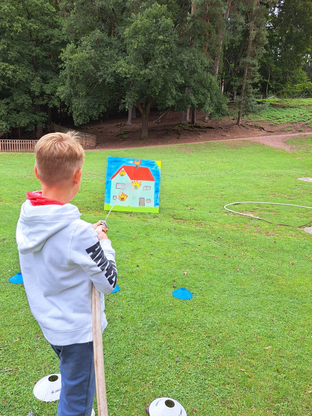 Ein Kind hält auf einer Wiese einen Feuerwehrschlauch und zielt mit einem Wasserstrahl auf eine Zielscheibe mit einem brennenden Haus. Die Aktion findet auf einer Freifläche vor einem Waldrand statt und vermittelt spielerisch den Umgang mit Löschtechnik.