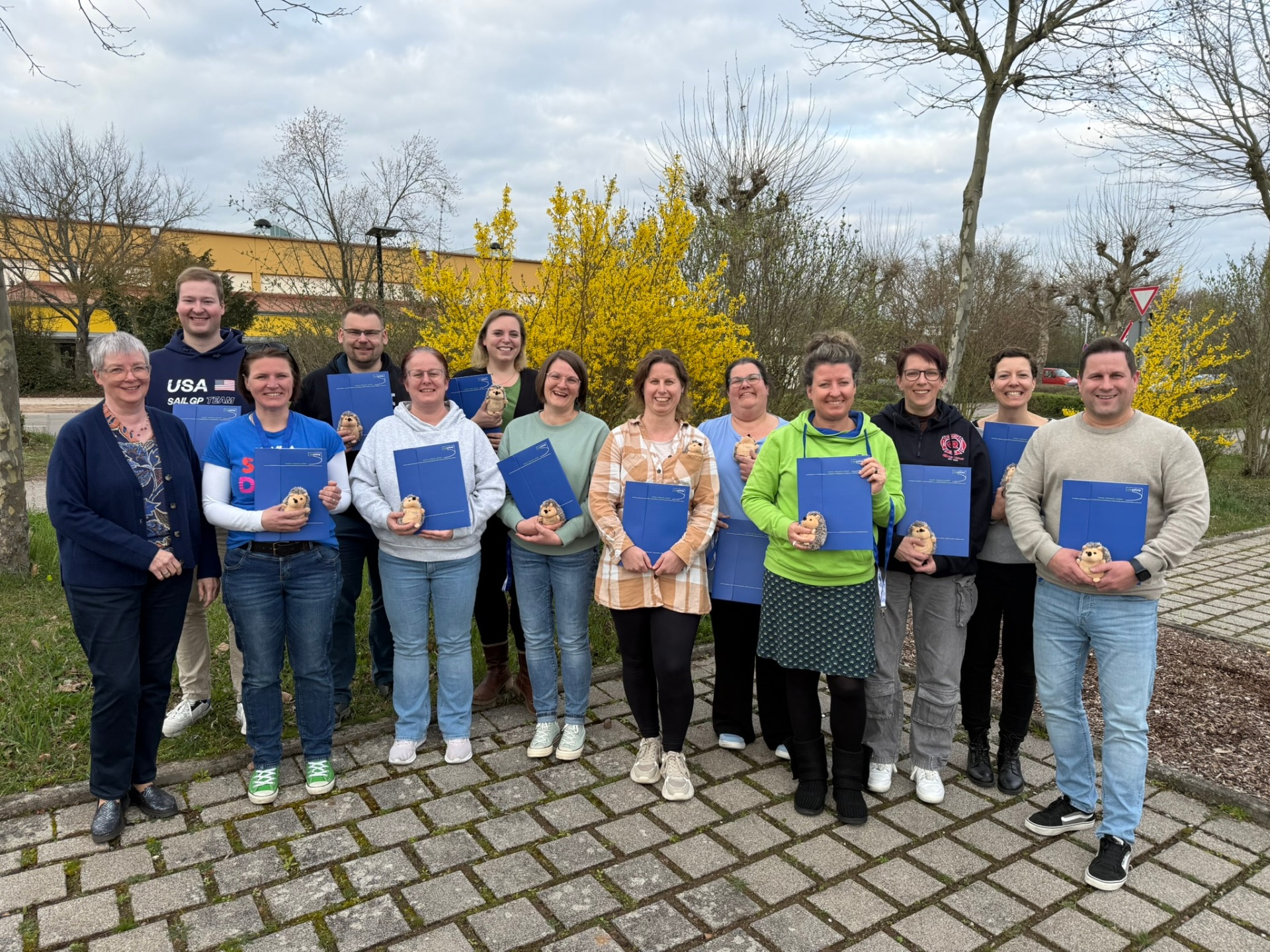 Gruppenfoto: 13 Personen des frisch ausgebildeten Einsatznachsorgeteams stehen im Freien auf einem Parkplatz. Alle halten eine blaue Mappe und eine kleinen Stoff-Igel in Händen. Im Hintergrund ist ein Grünstreifen mit teils blühenden Sträuchern zu sehen.