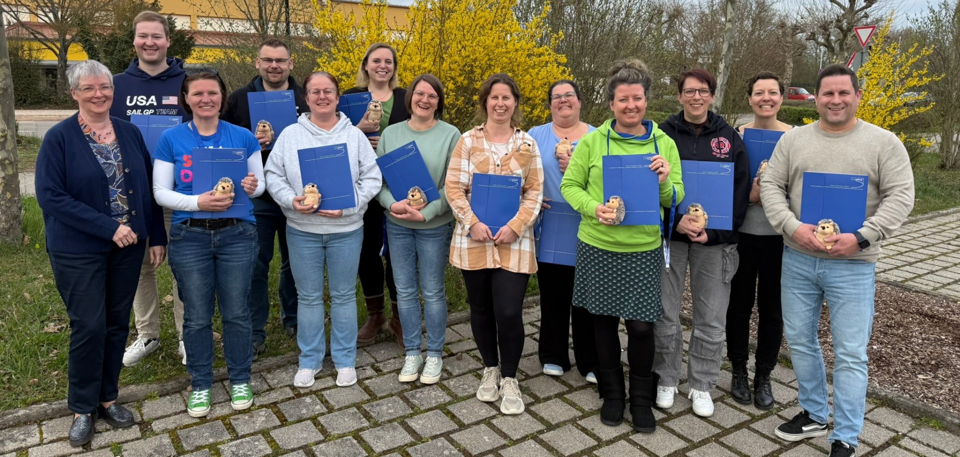 Gruppenfoto: 13 Personen des frisch ausgebildeten Einsatznachsorgeteams stehen im Freien auf einem Parkplatz. Alle halten eine blaue Mappe und eine kleinen Stoff-Igel in Händen. Im Hintergrund ist ein Grünstreifen mit teils blühenden Sträuchern zu sehen.