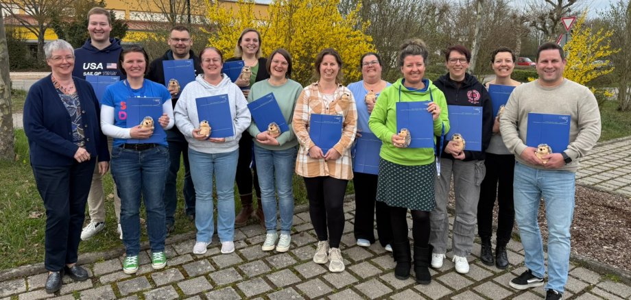 Gruppenfoto: 13 Personen des frisch ausgebildeten Einsatznachsorgeteams stehen im Freien auf einem Parkplatz. Alle halten eine blaue Mappe und eine kleinen Stoff-Igel in Händen. Im Hintergrund ist ein Grünstreifen mit teils blühenden Sträuchern zu sehen.