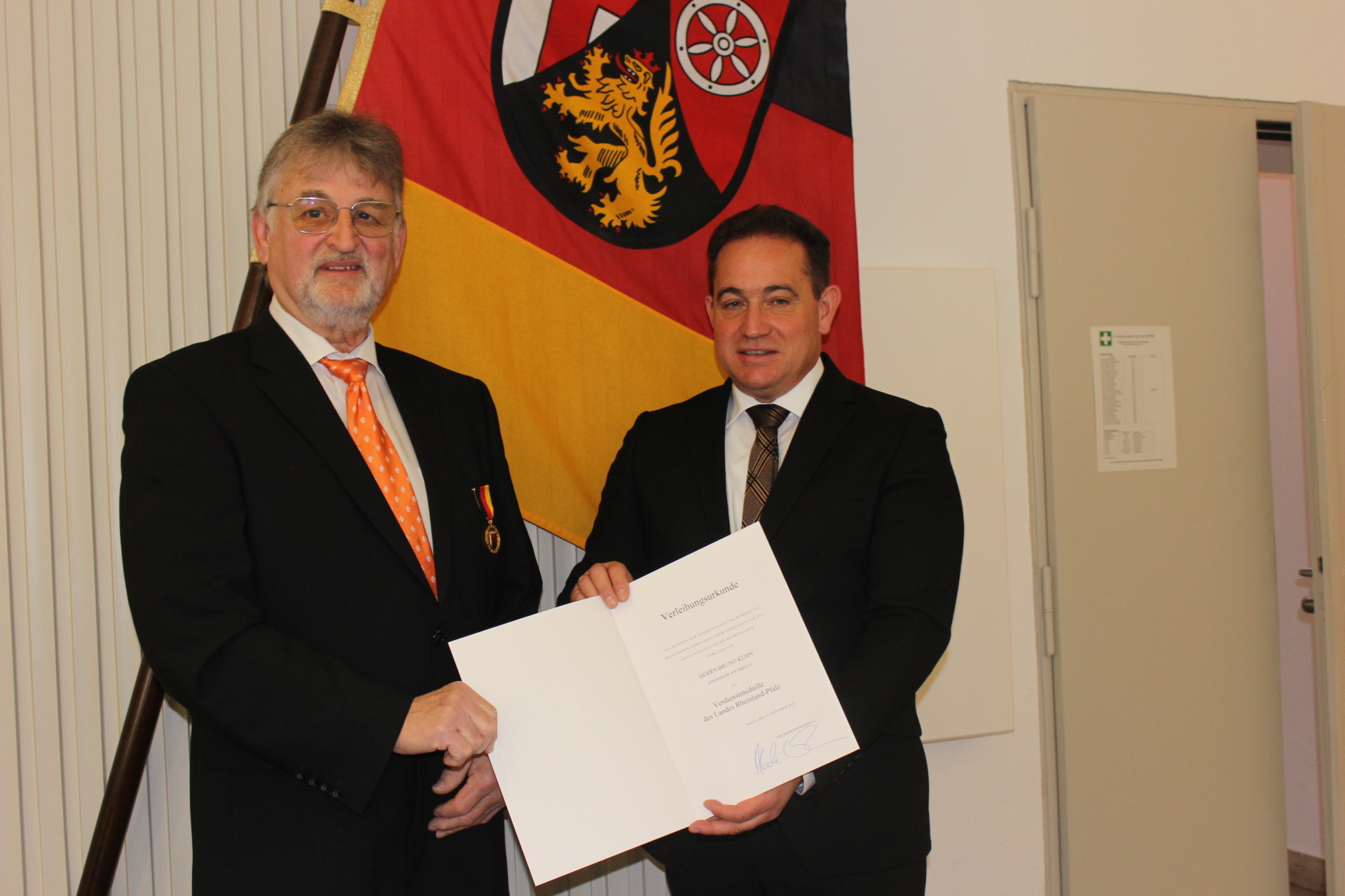 Zwei Männer in Anzügen stehen vor einer Rheinland-Pfalz-Flagge. Einer von ihnen hält eine geöffnete Verleihungsurkunde in der Hand, während der andere eine Medaille am Revers trägt.