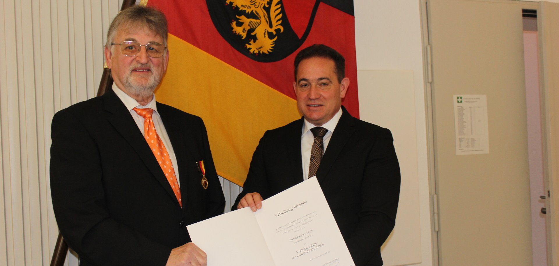 Zwei Männer in Anzügen stehen vor einer Rheinland-Pfalz-Flagge. Einer von ihnen hält eine geöffnete Verleihungsurkunde in der Hand, während der andere eine Medaille am Revers trägt.