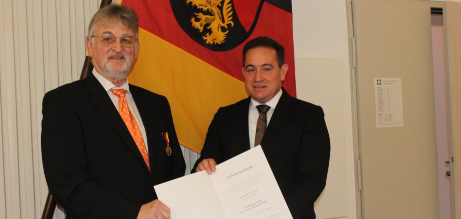 Zwei Männer in Anzügen stehen vor einer Rheinland-Pfalz-Flagge. Einer von ihnen hält eine geöffnete Verleihungsurkunde in der Hand, während der andere eine Medaille am Revers trägt.