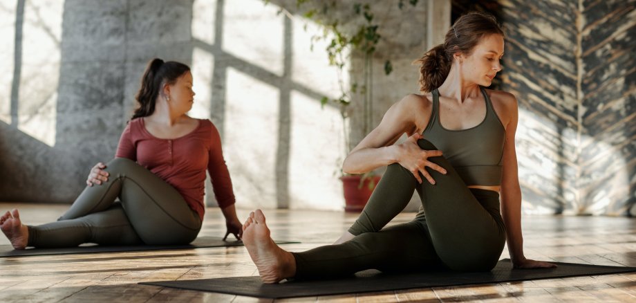 Zwei Frauen sitzen auf Yogamatten und machen Dehnungsübungen. 