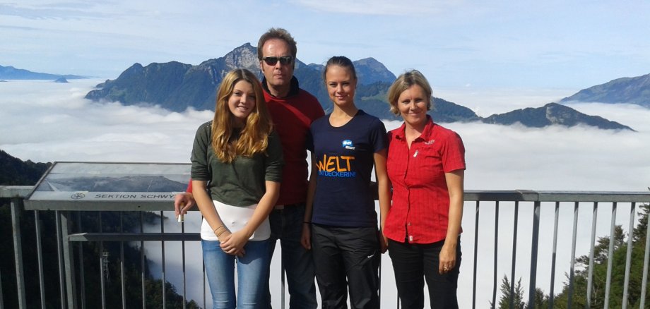Gruppenfoto einer Familie – Vater, Mutter, Tochter – mit einer Austauschschülerin. Die vier Personen stehen vor einem Geländer, hinter dem sich zunächst ein Tal und eine Berglandschaft erstrecken.