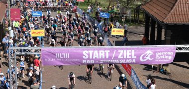 Zahlreiche Radfahrerinnen und Radfahrer starten bei einem Straßenrennen unter einem Banner mit der Aufschrift „Start / Ziel – Rad-Bundesliga“. Viele Zuschauerinnen und Zuschauer stehen entlang der Strecke und feuern das dicht gedrängte Fahrerfeld an.