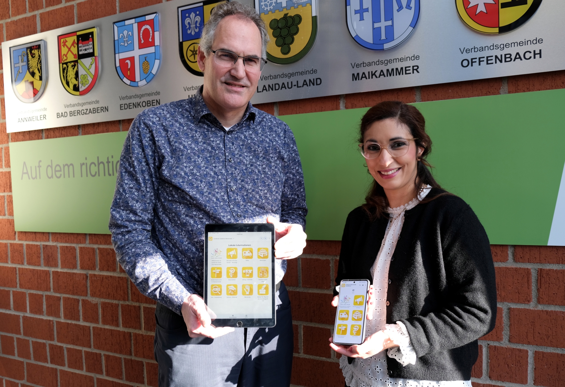 Landrat Dietmar Seefeldt und SÜW-Integrationsbeauftragte Sihame Hlubek stehen vor einer Wand mit den Wappen der Verbandsgemeinden des Landkreises Südliche Weinstraße. Beide halten digitale Geräte in die Kamera – ein Tablet und ein Smartphone – auf deren Bildschirmen eine App mit gelben Symbolen zu sehen ist.