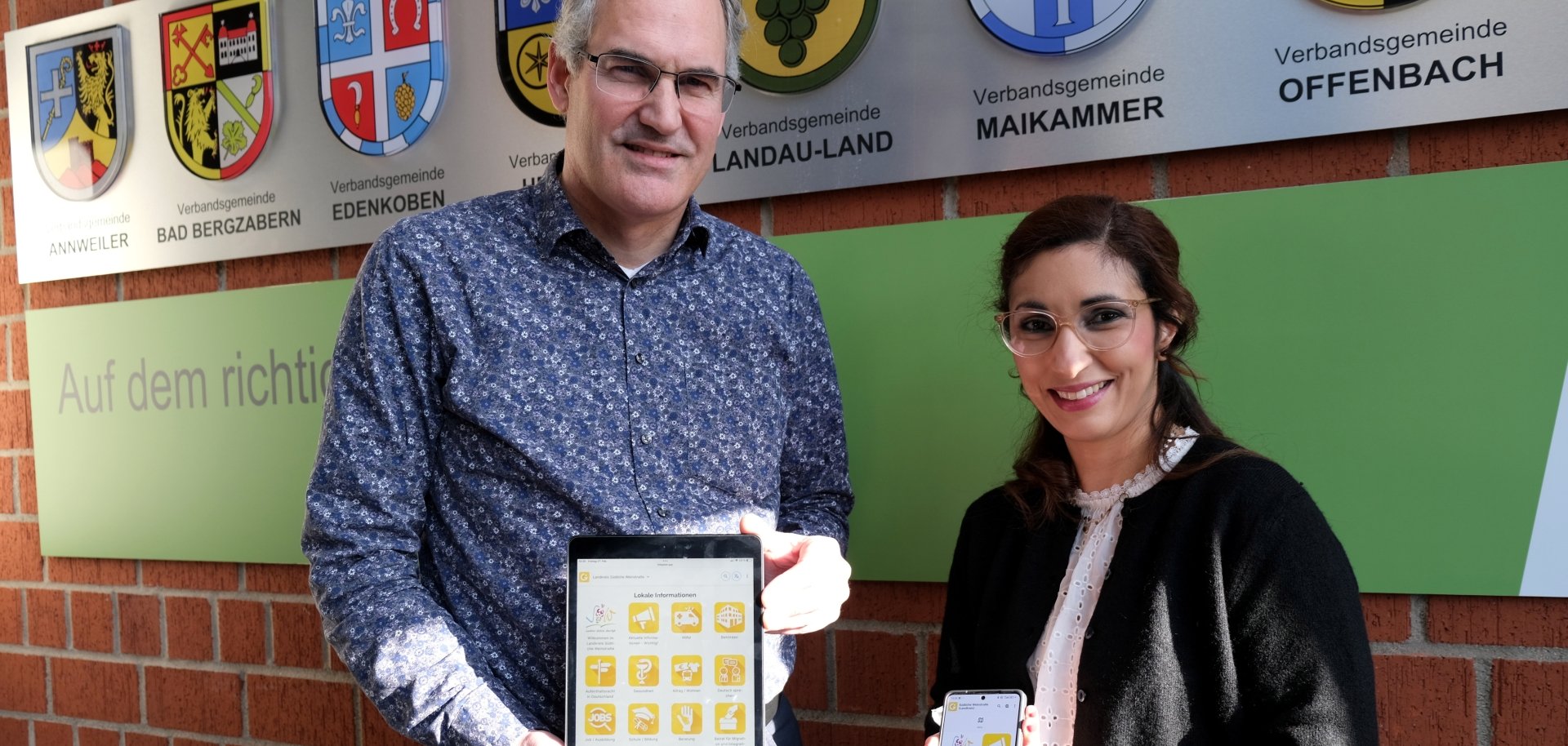 Landrat Dietmar Seefeldt und SÜW-Integrationsbeauftragte Sihame Hlubek stehen vor einer Wand mit den Wappen der Verbandsgemeinden des Landkreises Südliche Weinstraße. Beide halten digitale Geräte in die Kamera – ein Tablet und ein Smartphone – auf deren Bildschirmen eine App mit gelben Symbolen zu sehen ist.