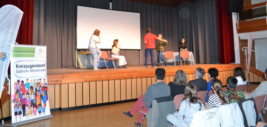 Eine Jugendtheatergruppe steht auf einer Bühne und spielt eine Szene vor Publikum. Im Vordergrund sitzen Zuschauerinnen und Zuschauer, links ist ein Banner des Kreisjugendamtes Südliche Weinstraße zu sehen.