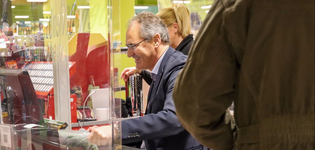 Landrat Dietmar Seefeldt zieht Waren über ein Kassenband einer Supermarkt-Kasse. Im Vordergrund rechts der Rücken eines Mannes, der gerade Einkäufe einpackt, im Hintergrund eine Supermarkt-Mitarbeiterin.
