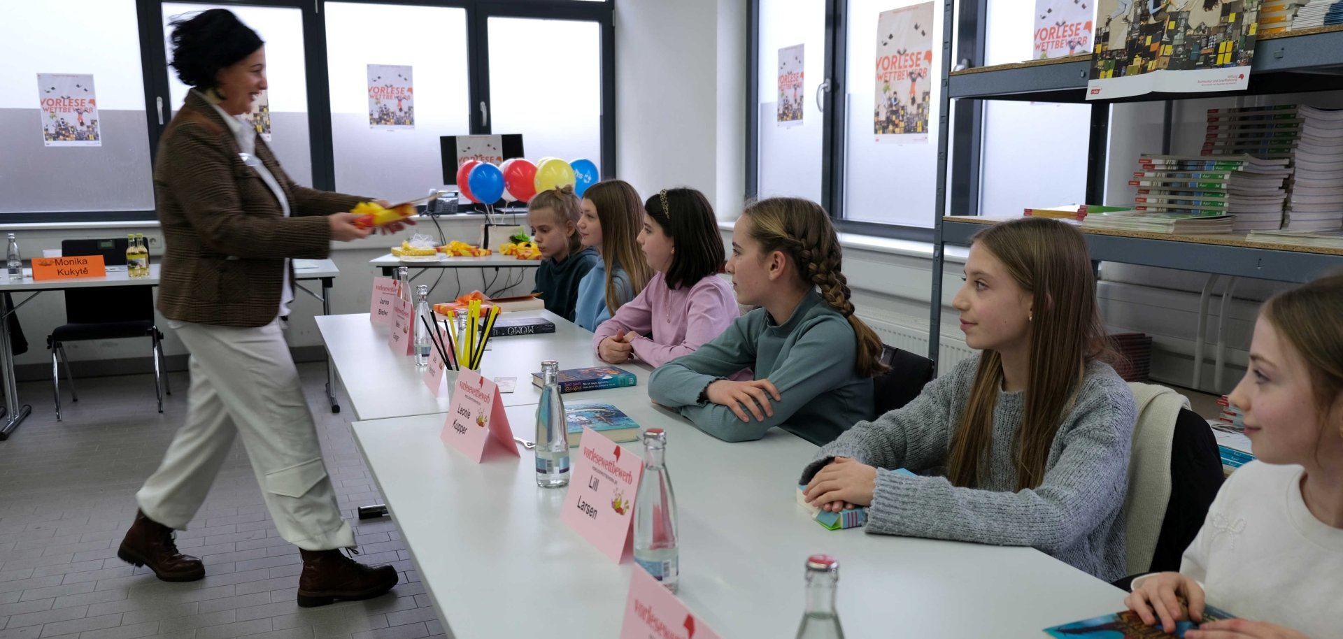 Mehrere Schülerinnen sitzen nebeneinander an einem Tisch mit Büchern, Namensschildern und Wasserflaschen. Eine Frau steht vor ihnen und überreicht ein Geschenk. An den Wänden hängen Plakate zum Vorlesewettbewerb, im Hintergrund sind Luftballons zu sehen.