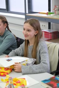 Lill Larsen heißt die Gewinnerin des diesjährigen Vorlesewettbewerbs Südliche Weinstraße. Foto: KV SÜW Eine Schülerin mit langen, offenen Haaren sitzt an einem Tisch und hält eine Urkunde auf einem Geschenk mit gelber Schleife. Sie lächelt leicht, vor ihr liegen Bücher und eine Wasserflasche.