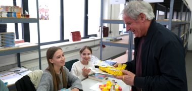 Kreisbeigeordneter Werner Schreiner überreicht Siegerin Lill Larsen in einem Klassenraum ein gelb verpacktes Geschenk. Das Mädchen sitzt an einem Tisch mit Büchern und einer Urkunde, eine weitere Schülerin daneben applaudiert. Im Hintergrund stehen Regale mit Schulbüchern.