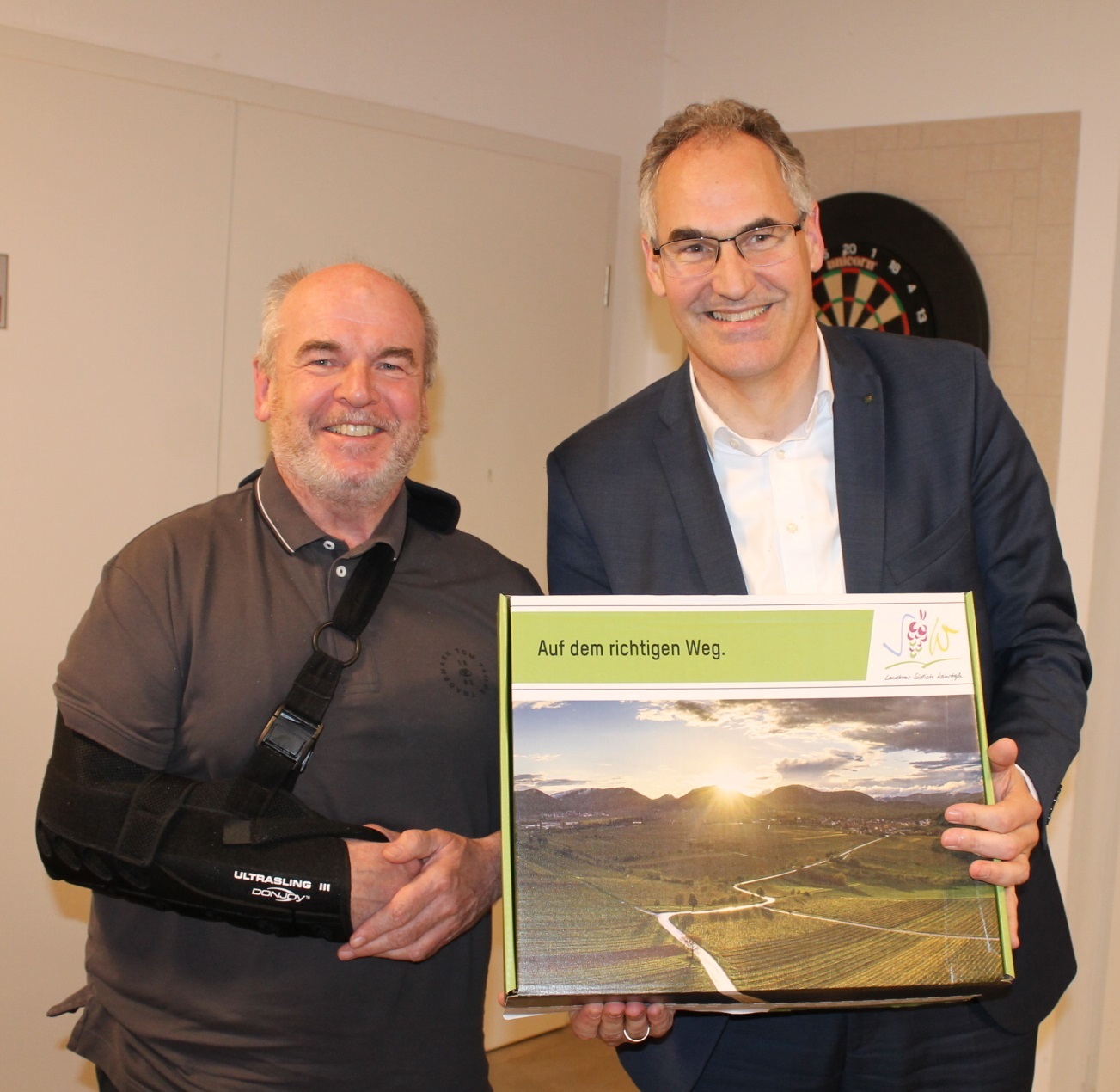 Zwei Männer stehen nebeneinander in einem Raum und lächeln. Einer trägt eine Armschlinge, der andere hält eine Präsentbox mit der Aufschrift „Auf dem richtigen Weg“ und einem Landschaftsbild in den Händen.