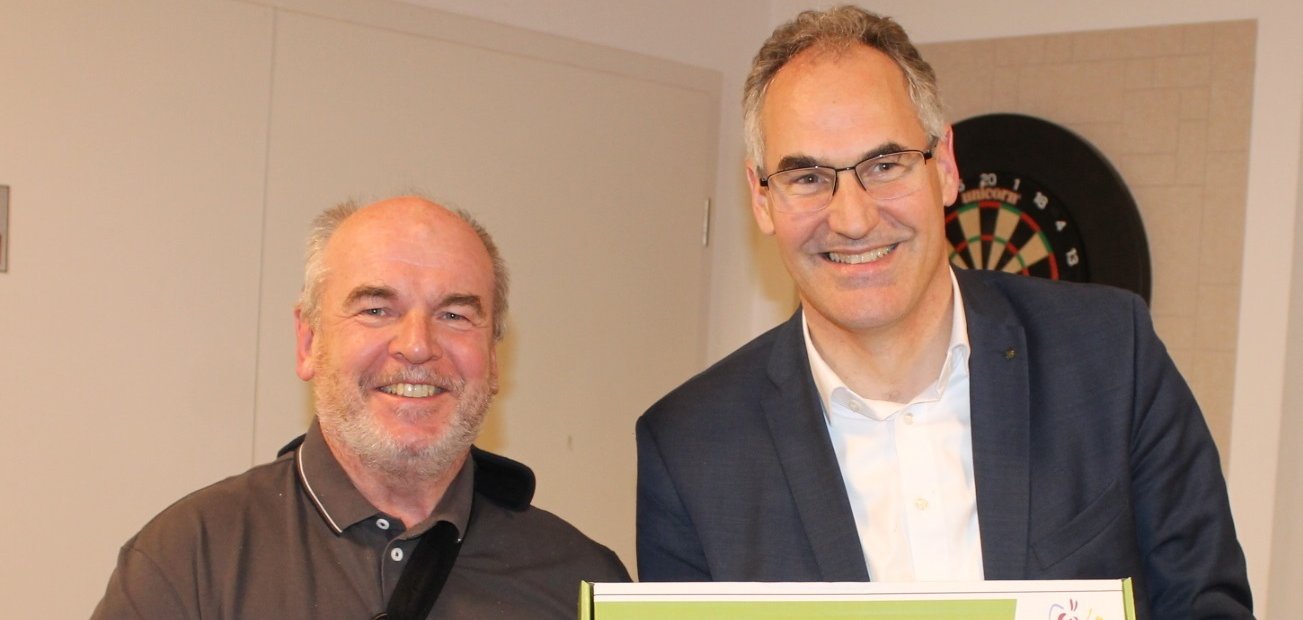 Zwei Männer stehen nebeneinander in einem Raum und lächeln. Einer trägt eine Armschlinge, der andere hält eine Präsentbox mit der Aufschrift „Auf dem richtigen Weg“ und einem Landschaftsbild in den Händen.