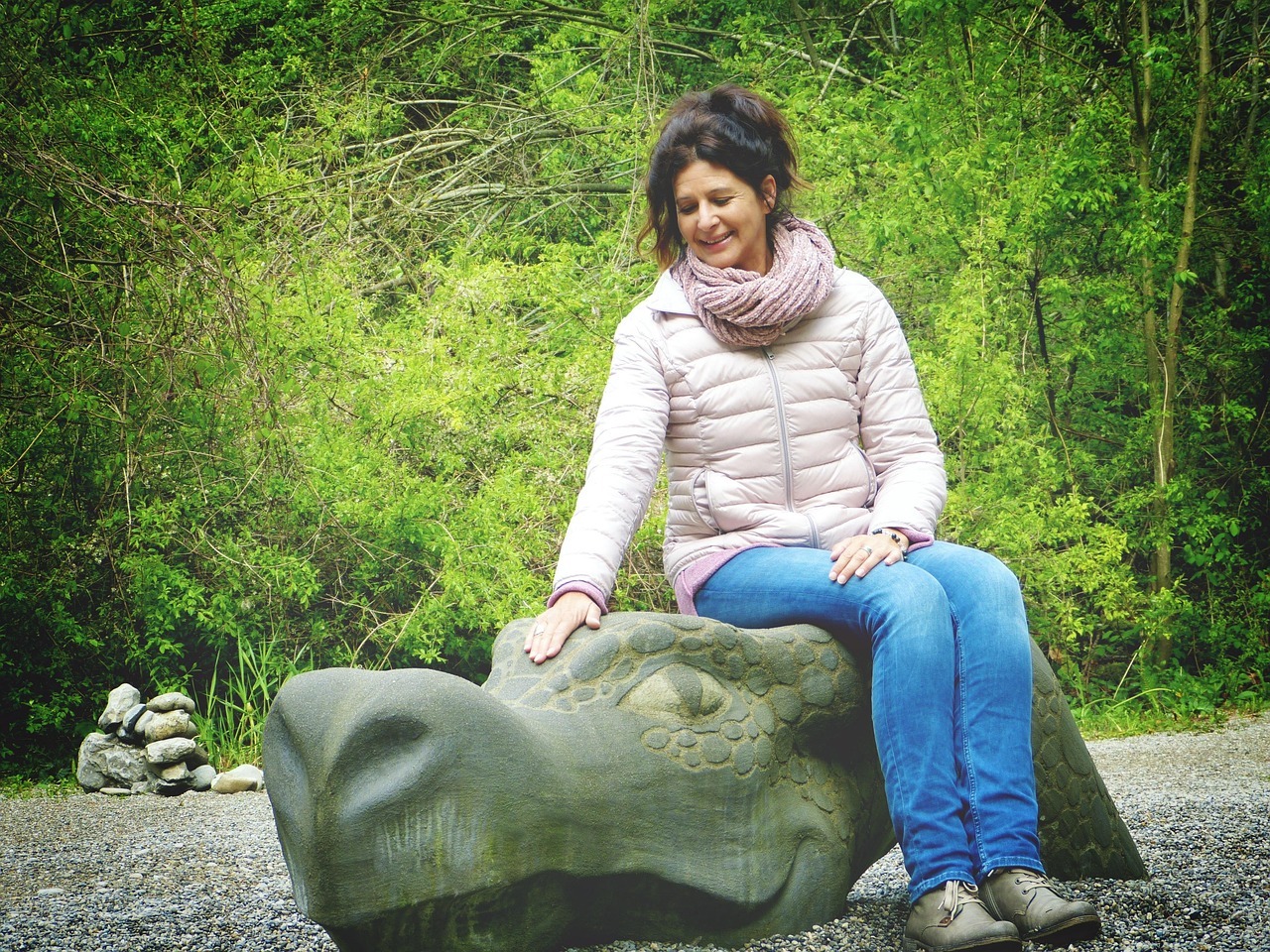 Frau im mittleren Alter sitzt im Freien auf einem steinernen Drachenkopf. Im Hintergrund sind grüne Bäume zu sehen.