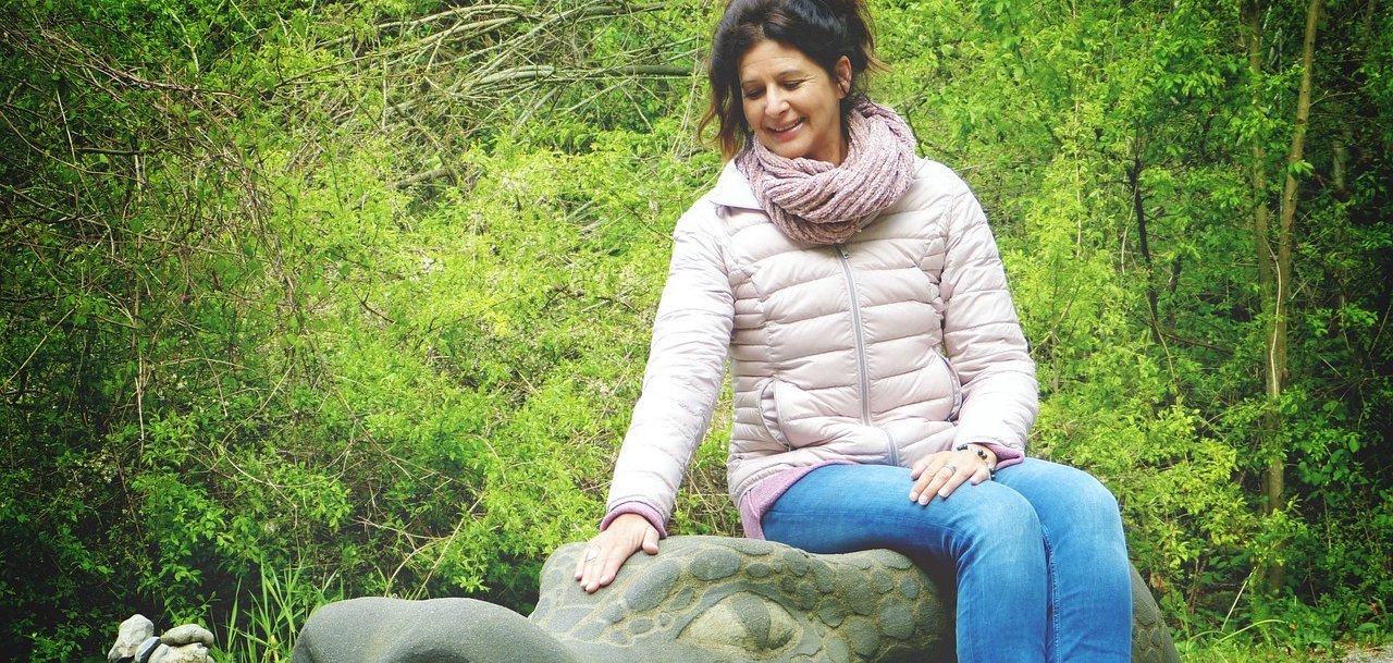 Frau im mittleren Alter sitzt im Freien auf einem steinernen Drachenkopf. Im Hintergrund sind grüne Bäume zu sehen.