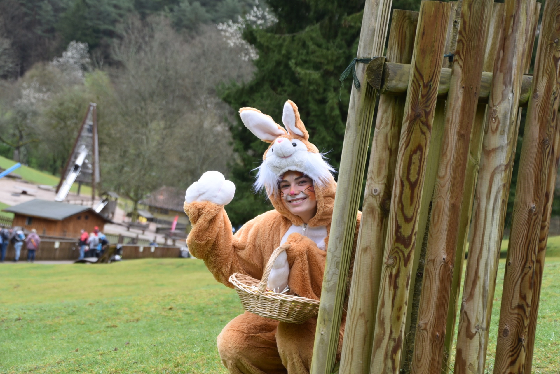 Eine als Osterhase verkleidete Person hockt auf einer Wiese hinter einem Holzpfosten, winkt freundlich und hält einen kleinen Korb in der Hand. Im Hintergrund sind ein Spielplatz und Bäume zu sehen.
