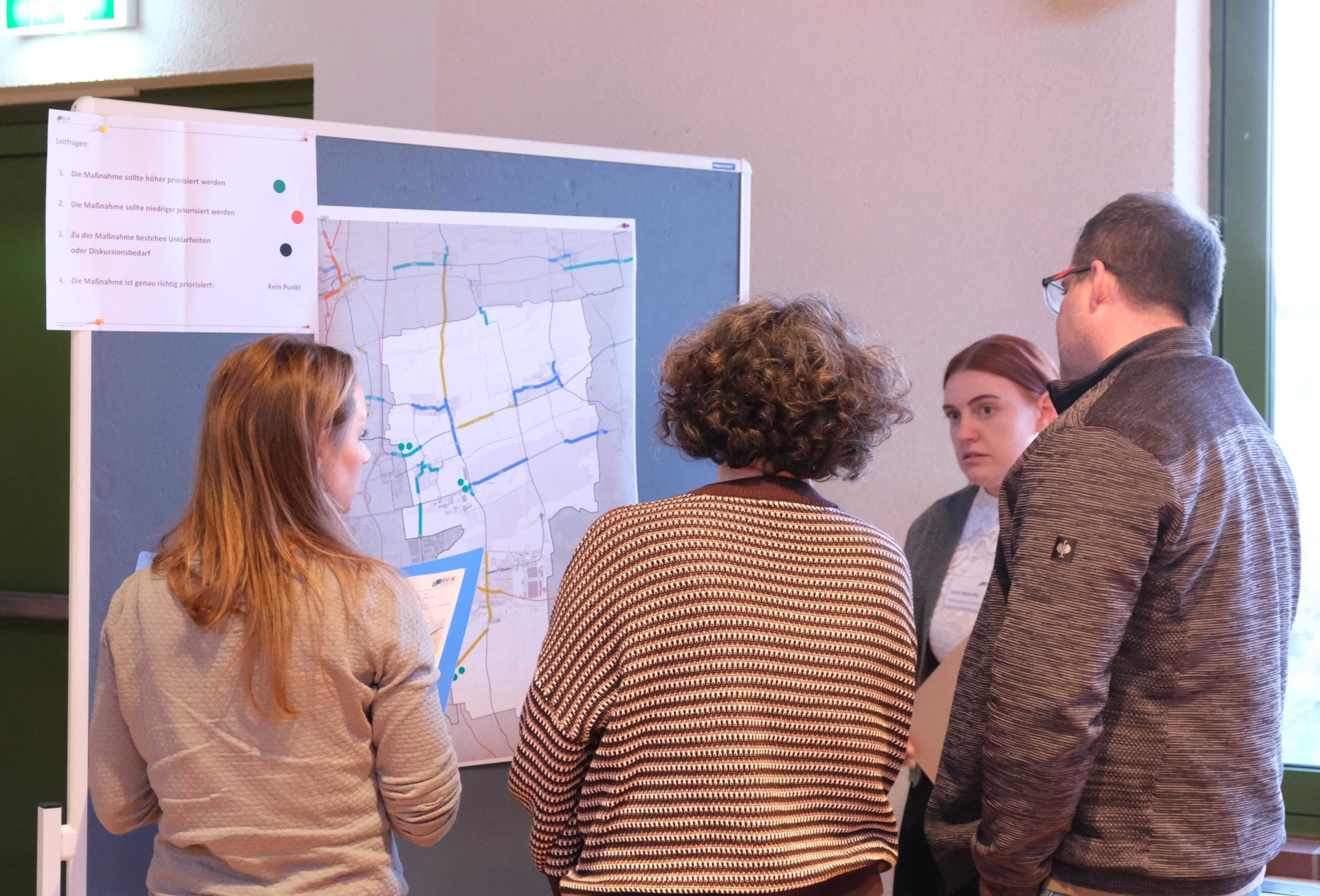 Vier Personen stehen vor einer Pinnwand mit einer großen Karte und diskutieren darüber. Auf der Karte sind farbig markierte Strecken und Punkte zu sehen, daneben hängt eine Legende mit Bewertungspunkten zur Priorisierung von Maßnahmen.