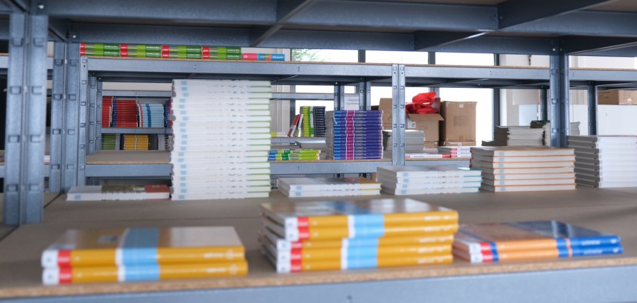 Gestapelte Schulbücher liegen auf einem Regal in einem Lagerraum. Im Hintergrund sind weitere Bücherregale mit Schulbüchern zu sehen.